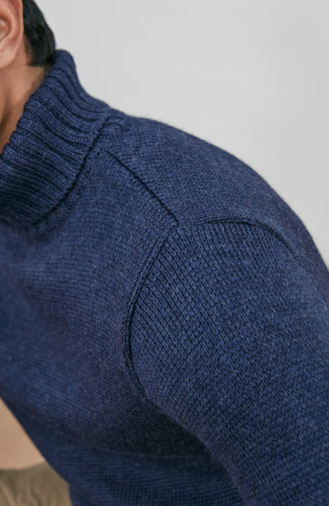A man wearing a navy blue knit wool turtleneck sweater made in england by paul james knitwear, a rust-orange ribbed merino wool beanie, and beige trousers, posed against a light gray studio background.