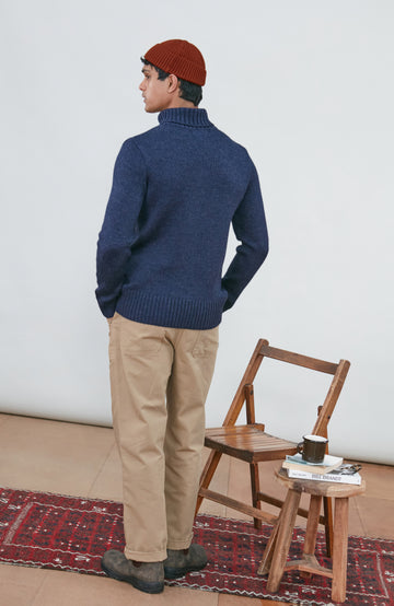 A man wearing a navy blue knit wool turtleneck sweater made in england by paul james knitwear, a rust-orange ribbed merino wool beanie, and beige trousers, posed against a light gray studio background.