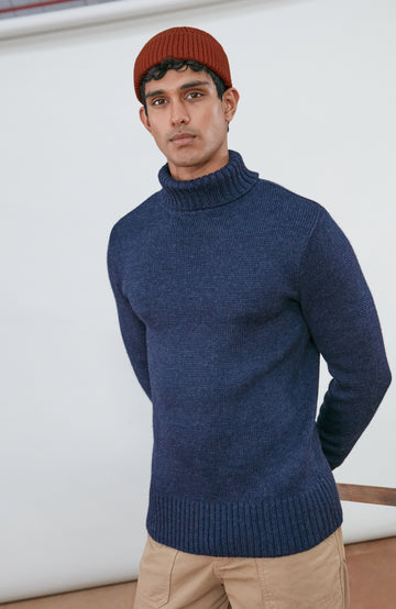 A man wearing a navy blue knit wool turtleneck sweater made in england by paul james knitwear, a rust-orange ribbed merino wool beanie, and beige trousers, posed against a light gray studio background.