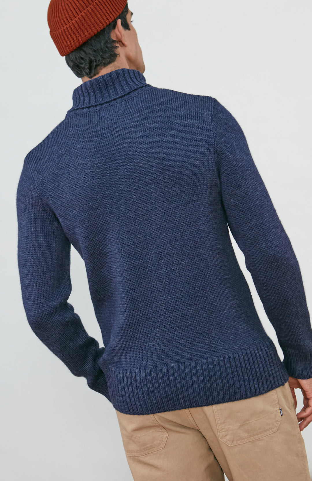 A man wearing a navy blue knit wool turtleneck sweater made in england by paul james knitwear, a rust-orange ribbed merino wool beanie, and beige trousers, posed against a light gray studio background.