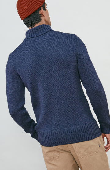 A man wearing a navy blue knit wool turtleneck sweater made in england by paul james knitwear, a rust-orange ribbed merino wool beanie, and beige trousers, posed against a light gray studio background.