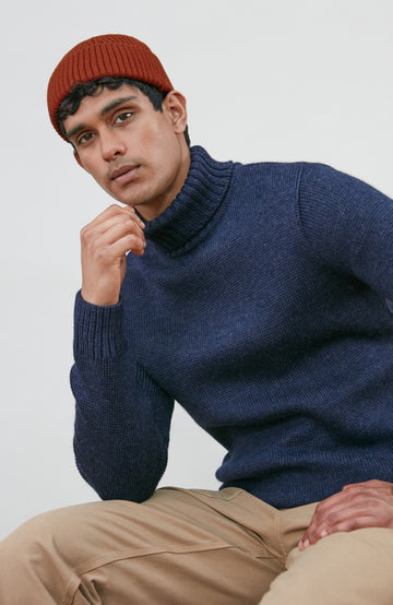 A man wearing a navy blue knit wool turtleneck sweater made in england by paul james knitwear, a rust-orange ribbed merino wool beanie, and beige trousers, posed against a light gray studio background.