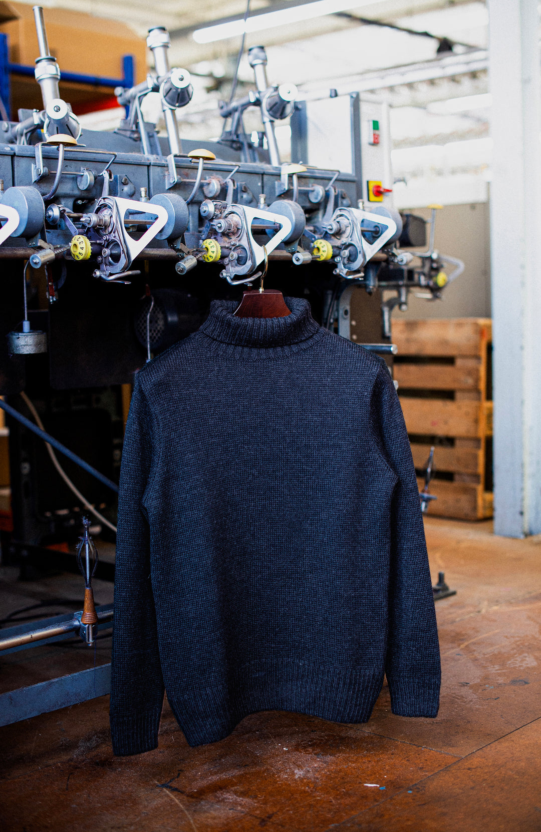 A dark charcoal gray knit turtleneck sweater hanging on a wooden hanger in front of a textile machinery machine in a workshop setting. In England men's wool sweater by Paul James Knitwear