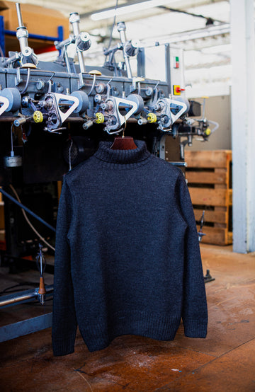 A dark charcoal gray knit turtleneck sweater hanging on a wooden hanger in front of a textile machinery machine in a workshop setting. In England men's wool sweater by Paul James Knitwear