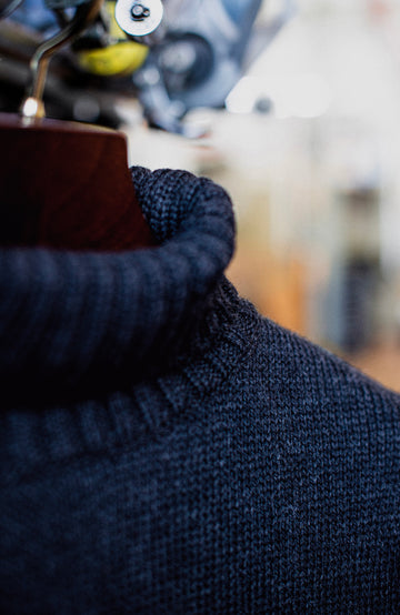 Close-up of a dark navy blue ribbed knit turtleneck sweater draped on a wooden hanger, with a softly blurred background. England Wool Sweater by Paul James Knitwear