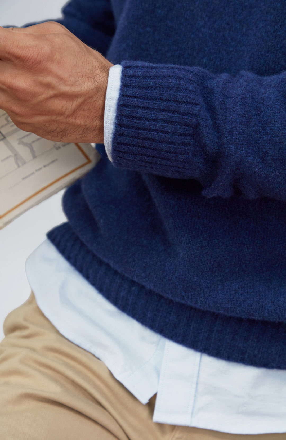 Made in England lambswool sweater by Paul James Knitwear. Close-up of a person wearing a navy blue knit sweater over a white collared shirt and beige trousers, with the hand adjusting the cuff.