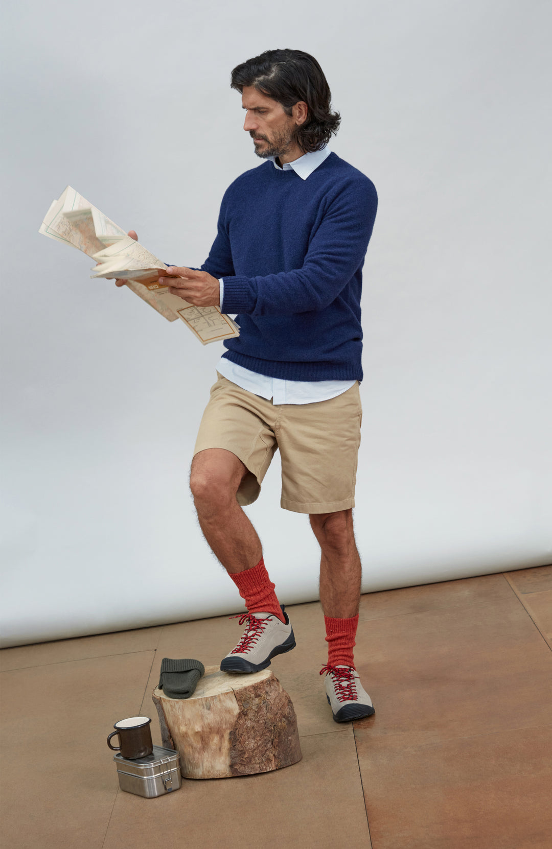 Made in England lambswool sweater by Paul James Knitwear. A man wearing a navy blue sweater, beige shorts, red socks, and hiking shoes stands on a small wooden stool, reading a map. The setting is a minimalist studio with a white backdrop and a brown floor.