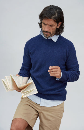 Made in England lambswool sweater by Paul James Knitwear. A man wearing a navy blue knit sweater over a light blue collared shirt and khaki shorts, holding an open book against a plain light gray studio background.