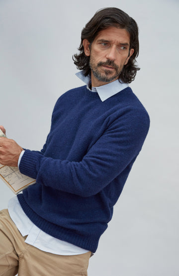Made in England lambswool sweater by Paul James Knitwear.  A man wearing a navy blue knit sweater over a light blue collared shirt and khaki pants, photographed against a plain light gray studio background.