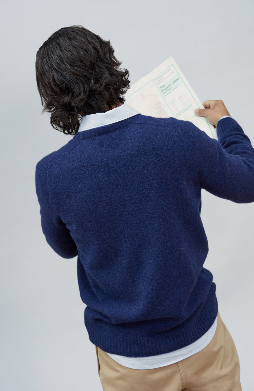 Made in England lambswool sweater by Paul James Knitwear. A man viewed from behind wearing a navy blue knit sweater over a white collared shirt and tan pants, holding a document against a light gray studio background.