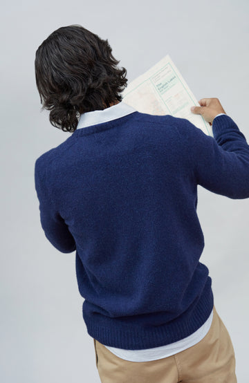 Made in England lambswool sweater by Paul James Knitwear. A man viewed from behind wearing a navy blue knit sweater over a white collared shirt and tan pants, holding a document against a light gray studio background.