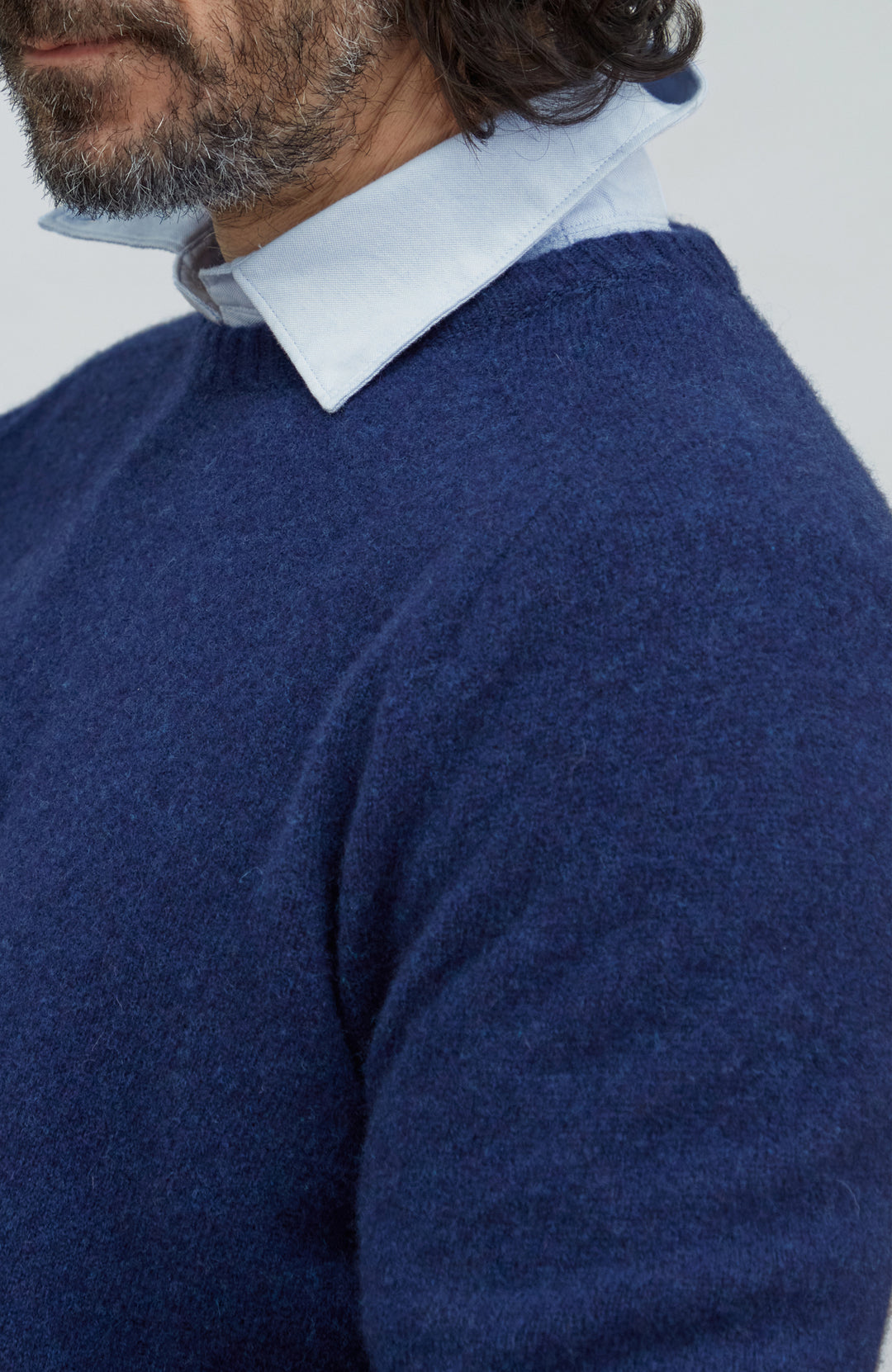 Made in England lambswool sweater by Paul James Knitwear. A close-up of a man wearing a navy blue knit sweater over a light blue collared shirt, shown from the chest up against a plain light gray background.