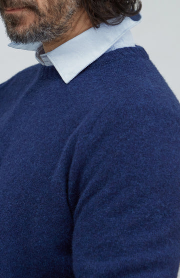 Made in England lambswool sweater by Paul James Knitwear. A close-up of a man wearing a navy blue knit sweater over a light blue collared shirt, shown from the chest up against a plain light gray background.