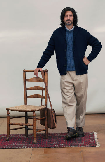 A man wearing a dark navy made in england merino wool navy cardigan by Paul James Knitwear over a textured blue knit wool fisherman sweater, beige trousers, and dark shoes, leaning on a wooden chair with a brown shoulder bag beside him.