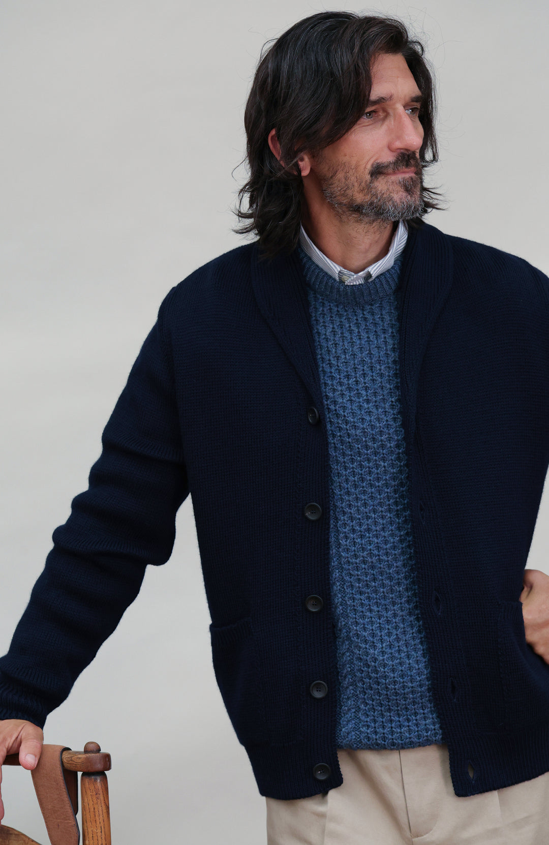 A man wearing a dark navy made in england merino wool navy cardigan by Paul James Knitwear over a textured blue knit wool fisherman sweater and beige trousers, standing against a light gray studio background.