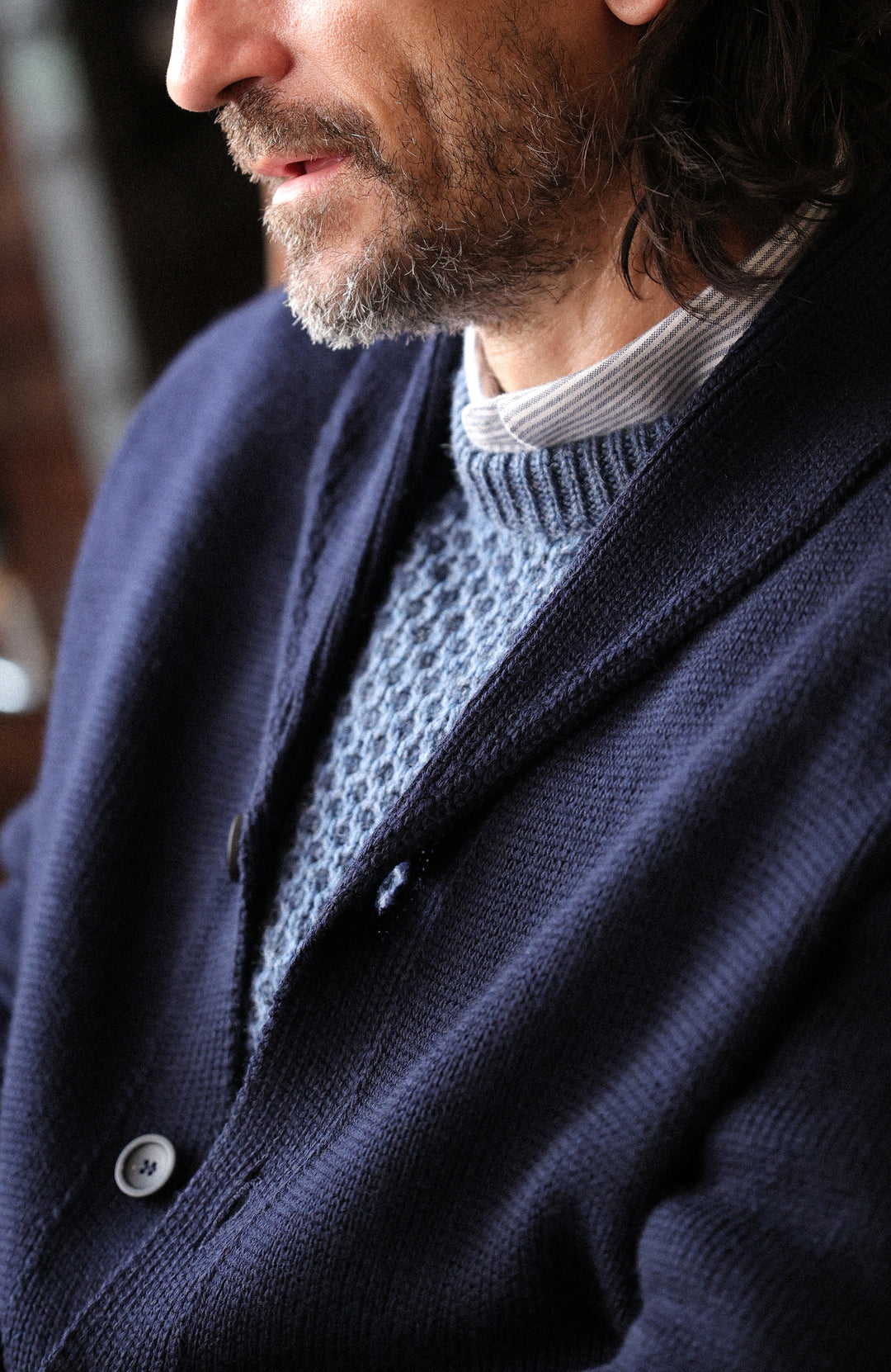 Chunky shawl-collar made-in-England cardigan by Paul James Knitwear. A close-up lifestyle shot of a man wearing a dark navy blue knit cardigan over a textured light blue-gray knit sweater.