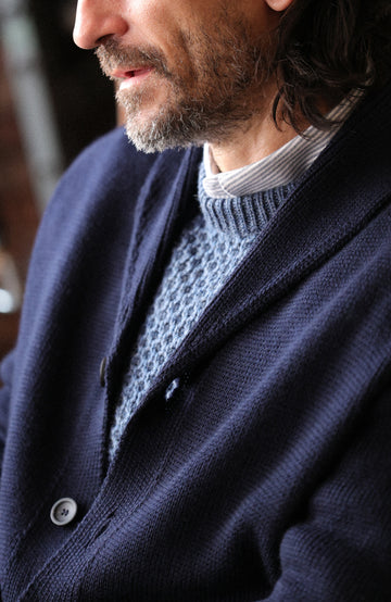 Chunky shawl-collar made-in-England cardigan by Paul James Knitwear. A close-up lifestyle shot of a man wearing a dark navy blue knit cardigan over a textured light blue-gray knit sweater.