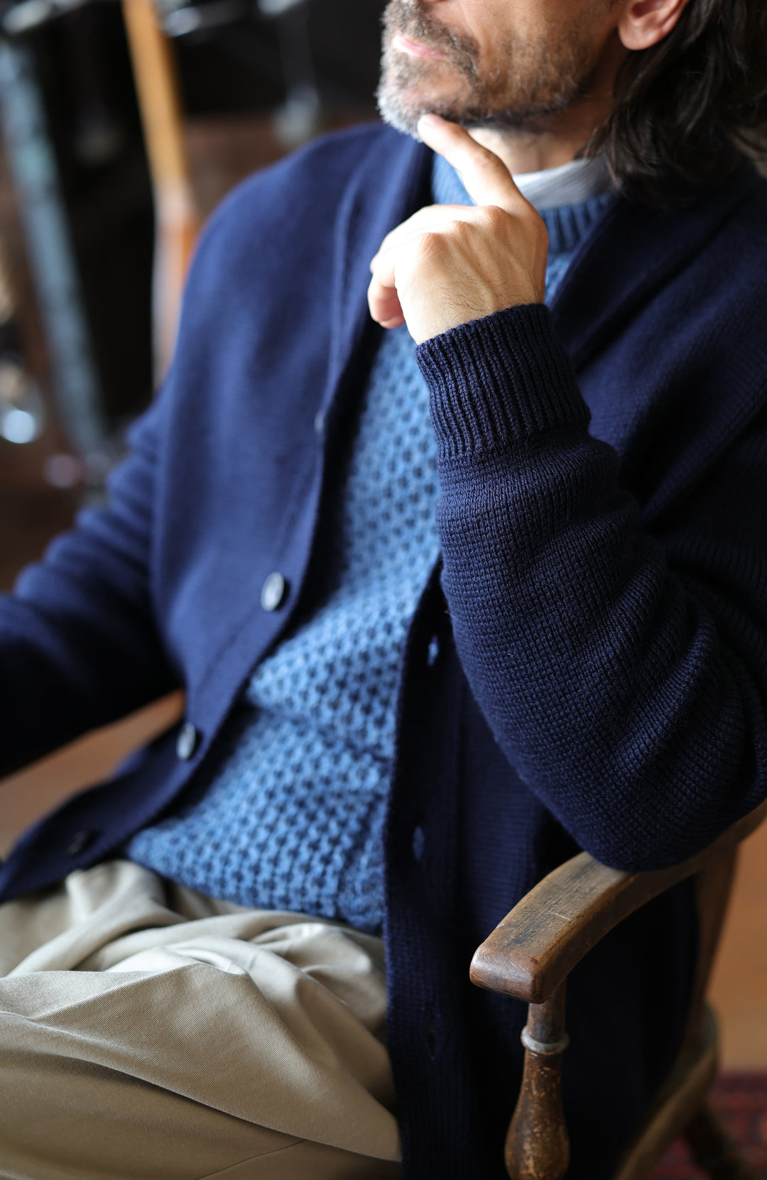 Chunky shawl-collar made-in-England cardigan by Paul James Knitwear. A man wearing a navy blue knit cardigan over a textured blue sweater and beige trousers, sitting in a wooden chair in a dimly lit indoor setting.