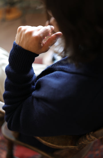 Chunky shawl-collar made-in-England cardigan by Paul James Knitwear. A close-up lifestyle shot of a person wearing a dark navy blue ribbed knit sweater, seen from behind as they sit on a wooden chair.
