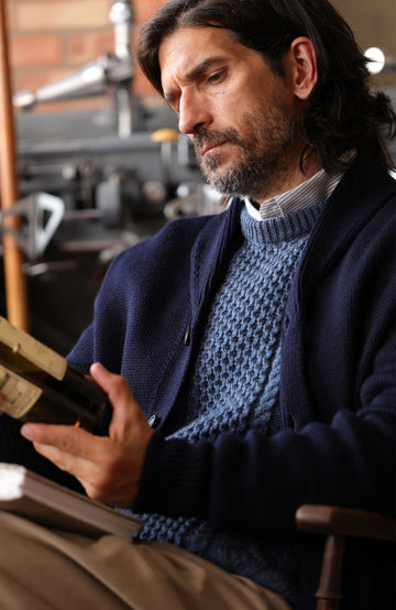 A man wearing a dark navy made in england merino wool navy shawl collar cardigan by Paul James Knitwear over a textured blue knit wool fisherman sweater
