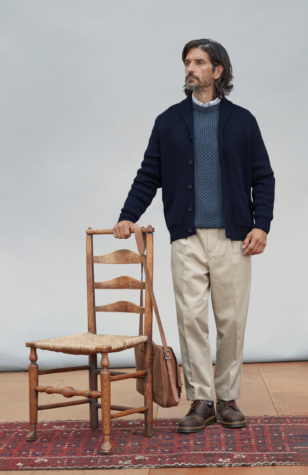 A man stands in a studio wearing a navy cardigan over a blue patterned sweater, with beige trousers and brown shoes. He is holding the back of a wooden chair and carrying a brown shoulder bag, standing on a red patterned rug against a light gray background. Chunky shawl-collar made-in-England cardigan by Paul James Knitwear