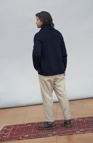 Chunky shawl-collar made-in-England cardigan by Paul James Knitwear. A man stands with his back to the camera wearing a dark navy jacket, beige trousers, and brown shoes, standing on a red patterned rug against a light gray studio backdrop.