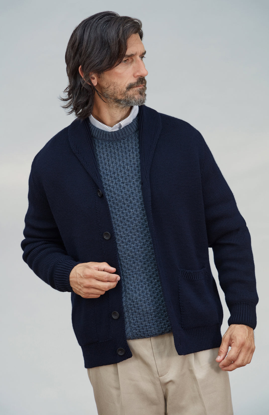 A man wearing a navy blue knit cardigan over a textured blue sweater, a white collared shirt, and beige trousers, photographed against a light gray studio background. Chunky short-collar made-in-England cardigan by Paul James Knitwear