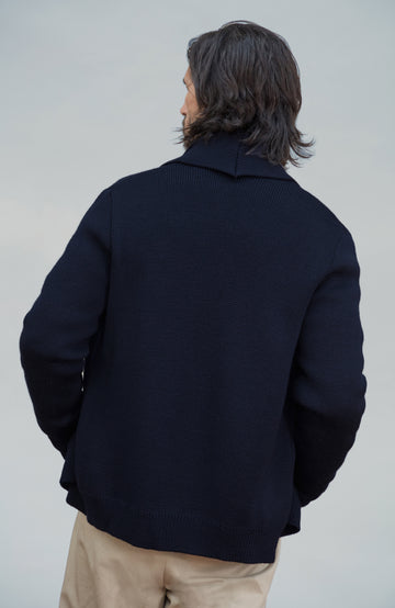 Chunky shawl-collar made-in-England cardigan by Paul James Knitwear. A man with long dark hair is shown from behind wearing a dark navy knit cardigan sweater and beige trousers, photographed against a plain light gray studio background.