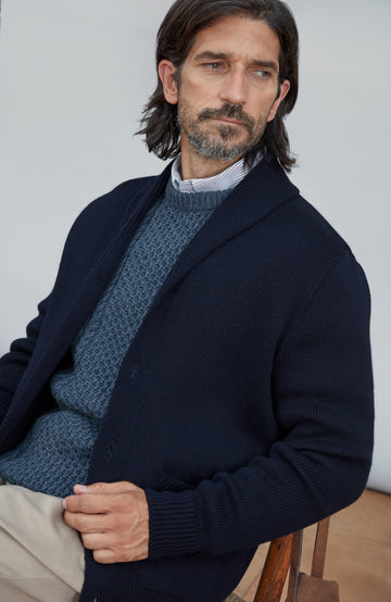 A man wearing a dark navy blue knit cardigan over a textured blue sweater, a striped shirt, and beige trousers, seated against a light gray background. Chunky shawl-collar made-in-England cardigan by Paul James Knitwear
