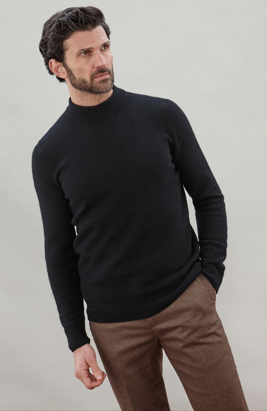 A man wearing a black long-sleeve wool mock neck made in England sweater and brown trousers, standing against a neutral light gray background.