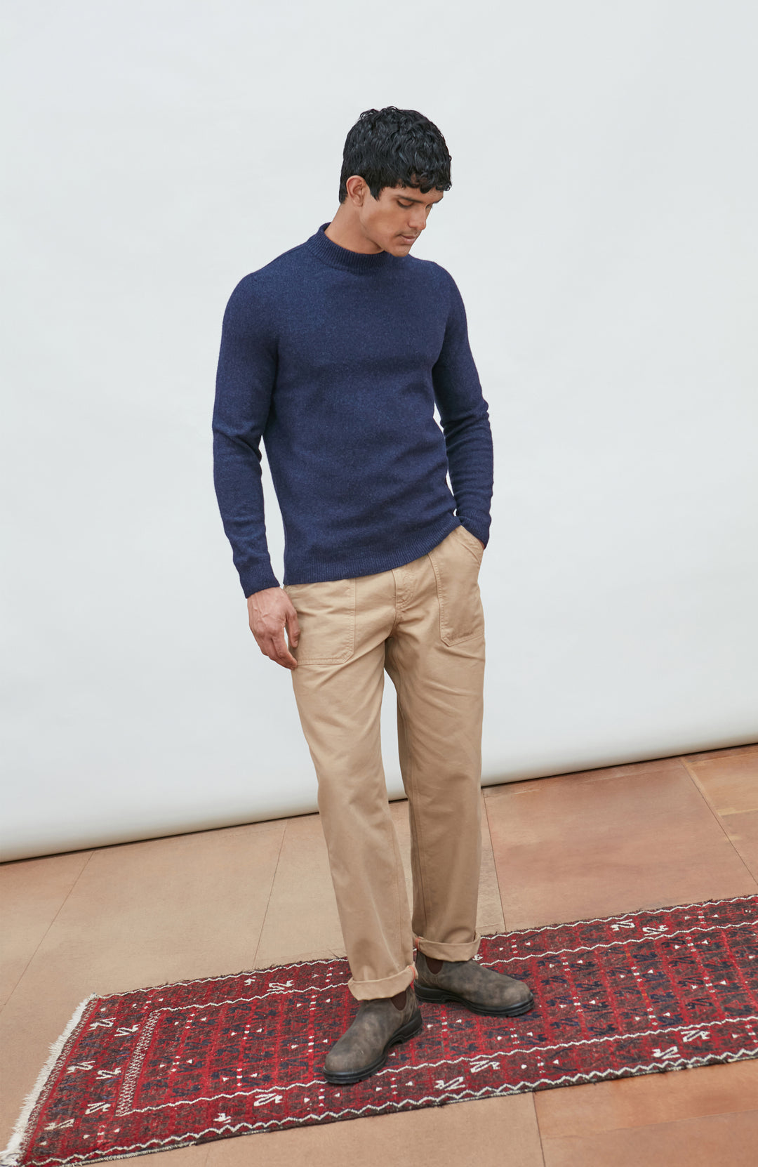 A man wearing a navy blue knit mock neck wool sweater by paul james knitwear made in england and khaki pants, photographed against a light gray studio background.