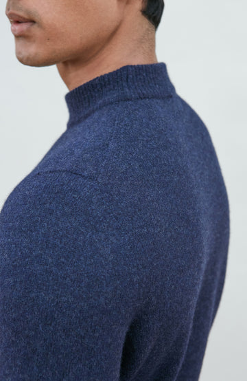 A man wearing a navy blue knit mock neck wool sweater by paul james knitwear made in england and khaki pants, photographed against a light gray studio background.