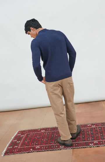 A man wearing a navy blue knit mock neck wool sweater by paul james knitwear made in england and khaki pants, photographed against a light gray studio background.