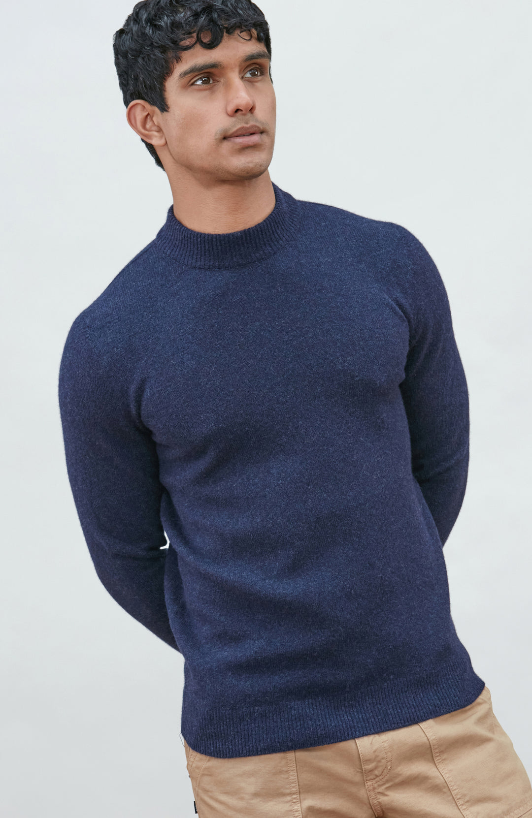 A man wearing a navy blue knit mock neck wool sweater by paul james knitwear made in england and khaki pants, photographed against a light gray studio background.
