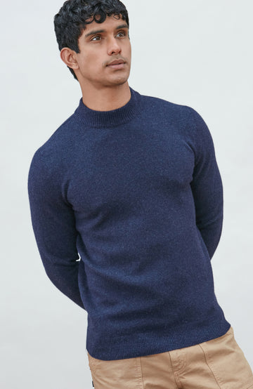 A man wearing a navy blue knit mock neck wool sweater by paul james knitwear made in england and khaki pants, photographed against a light gray studio background.