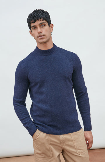 A man wearing a navy blue knit mock neck wool sweater by paul james knitwear made in england and khaki pants, photographed against a light gray studio background.
