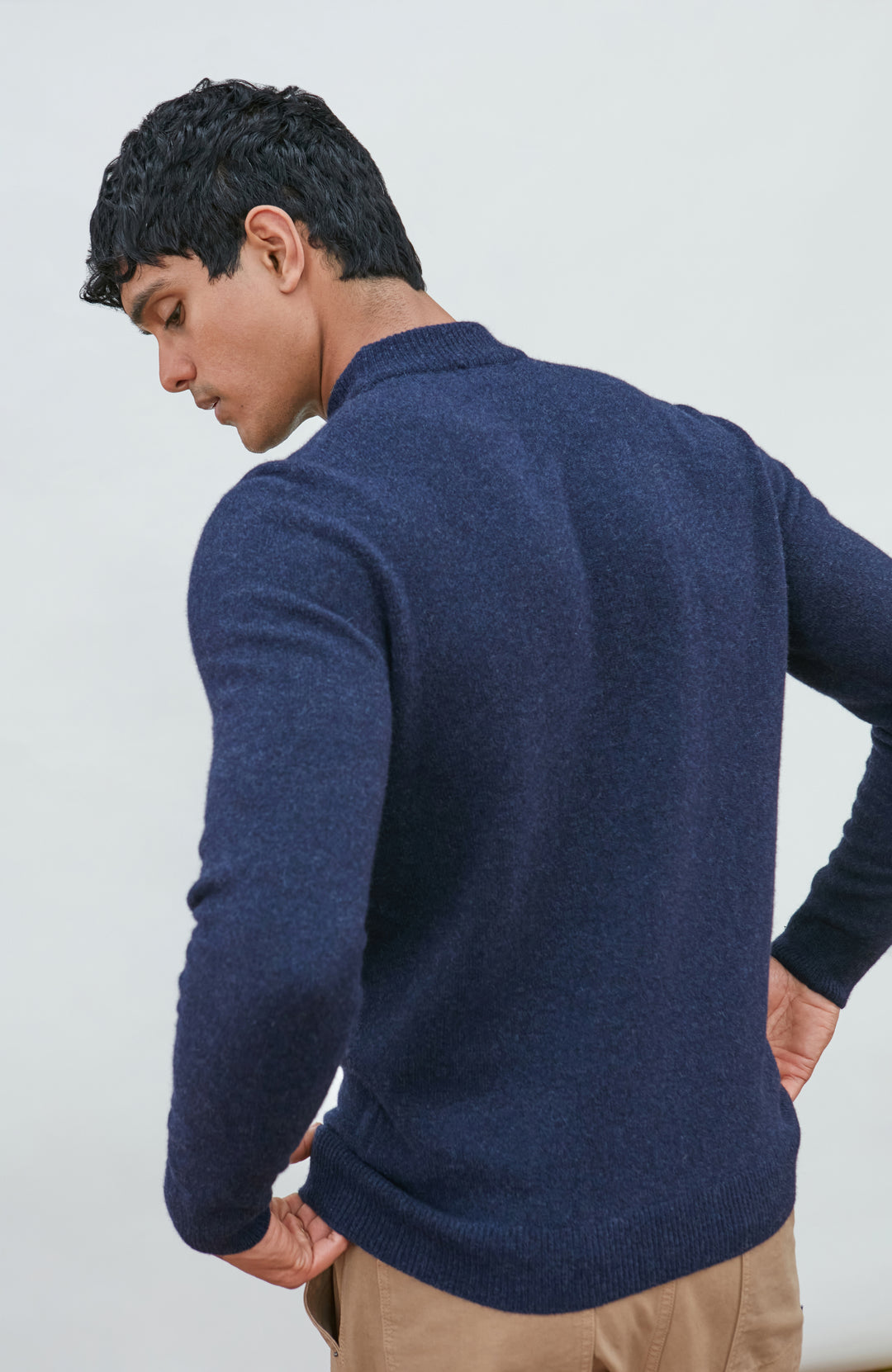A man wearing a navy blue knit mock neck wool sweater by paul james knitwear made in england and khaki pants, photographed against a light gray studio background.