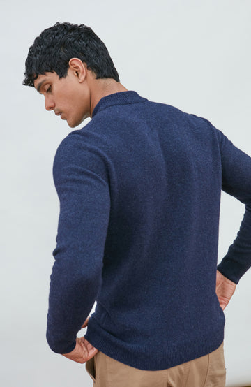 A man wearing a navy blue knit mock neck wool sweater by paul james knitwear made in england and khaki pants, photographed against a light gray studio background.
