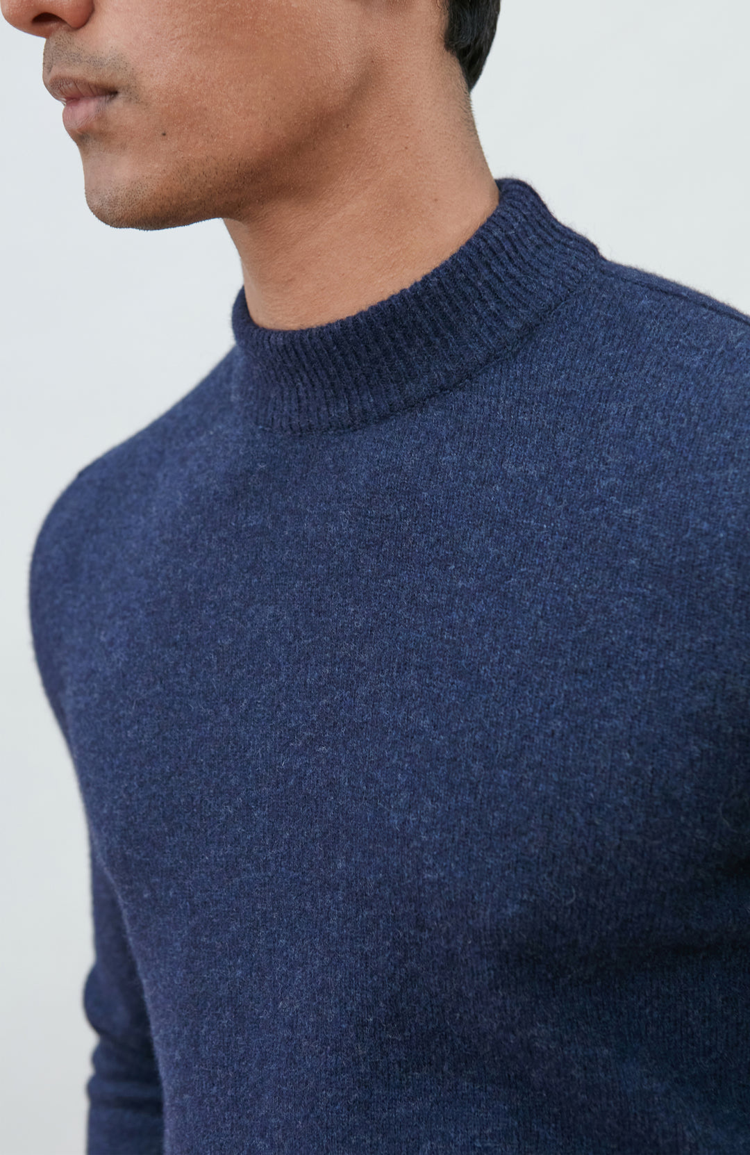 A man wearing a navy blue knit mock neck wool sweater by paul james knitwear made in england and khaki pants, photographed against a light gray studio background.