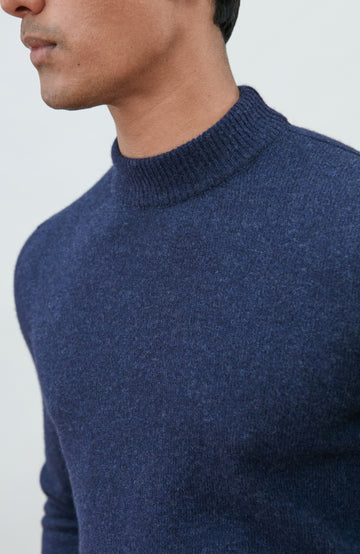 A man wearing a navy blue knit mock neck wool sweater by paul james knitwear made in england and khaki pants, photographed against a light gray studio background.
