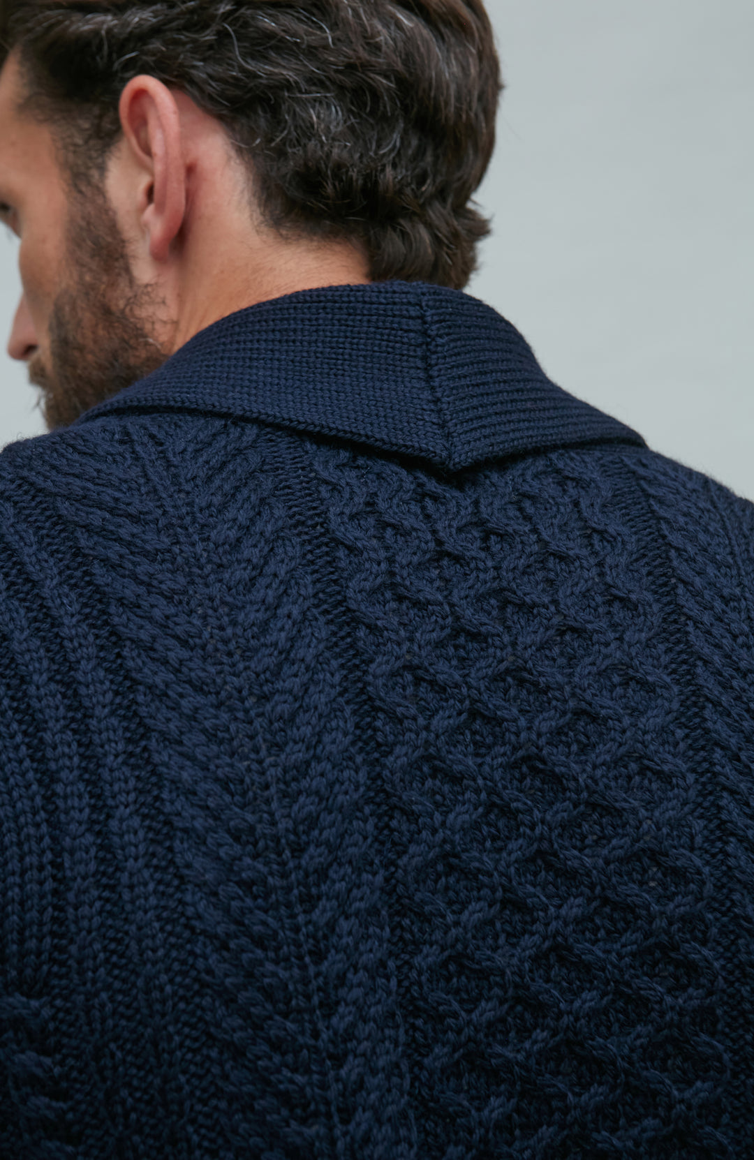 Man wearing a navy wool cable knit cardigan sitting on a wooden chair by Paul James