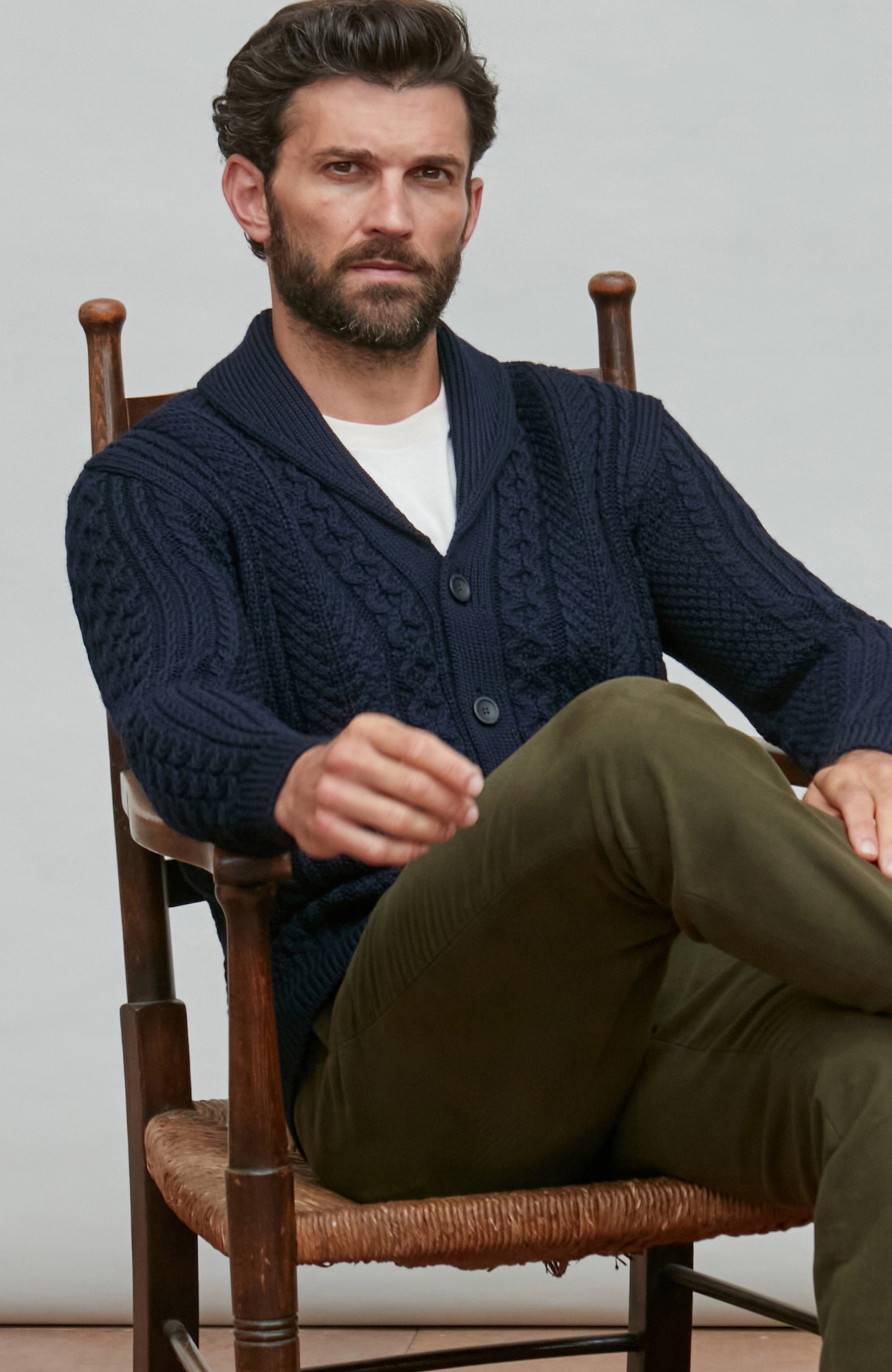 Man wearing a navy wool cable knit cardigan sitting on a wooden chair.