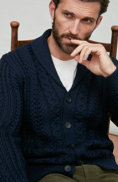 Man wearing a navy wool cable knit cardigan sitting on a wooden chair.