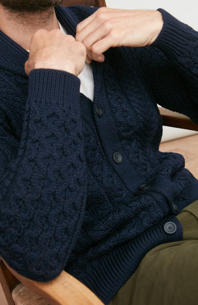 Man wearing a navy wool cable knit cardigan sitting on a wooden chair.