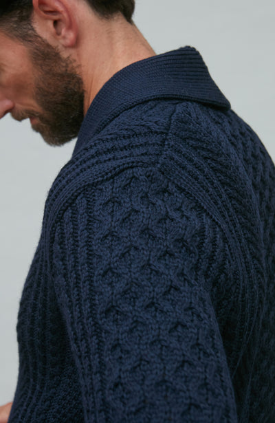 Man wearing a navy wool cable knit cardigan sitting on a wooden chair.