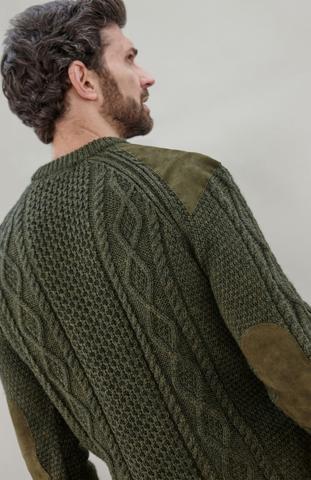 A man wearing an olive green cable-knit sweater with brown suede elbow patches, shown from a side-back angle against a plain light gray background.