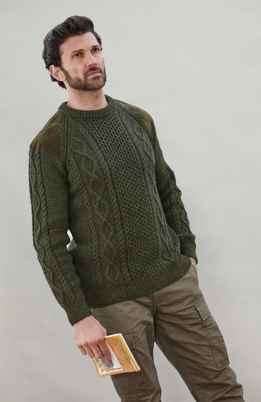 A man wearing a dark olive green cable-knit made in England sweater by Paul James Knitwear with contrasting solid green sleeves, paired with olive cargo pants, standing against a neutral light gray background.