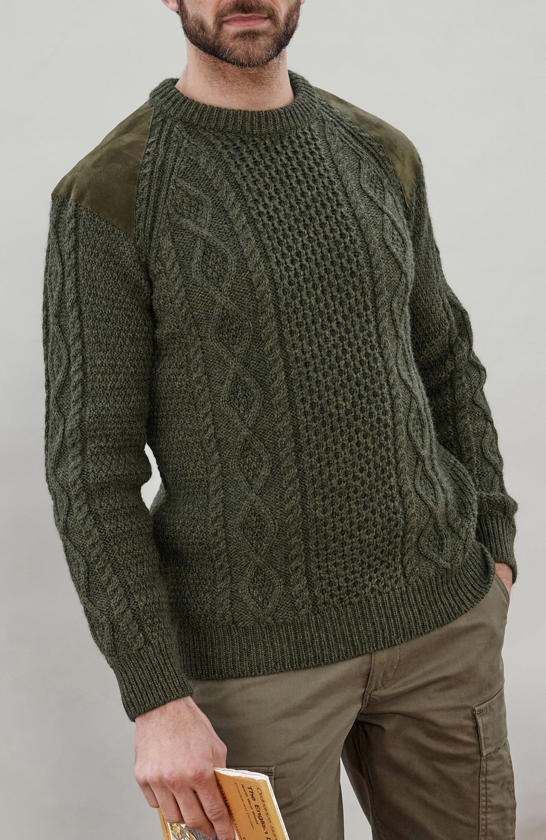 A man wearing an olive green cable-knit sweater with contrasting solid knit sleeves and olive green shoulder patches, paired with khaki cargo pants against a neutral gray background.
