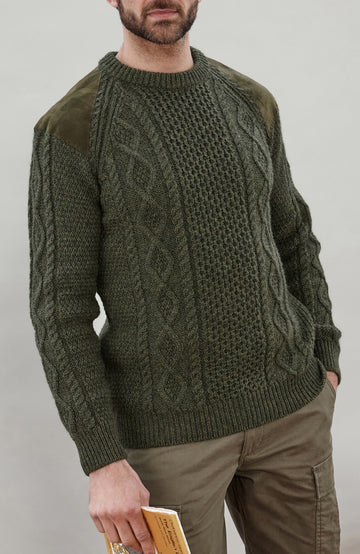 A man wearing an olive green cable-knit sweater with contrasting solid knit sleeves and olive green shoulder patches, paired with khaki cargo pants against a neutral gray background.