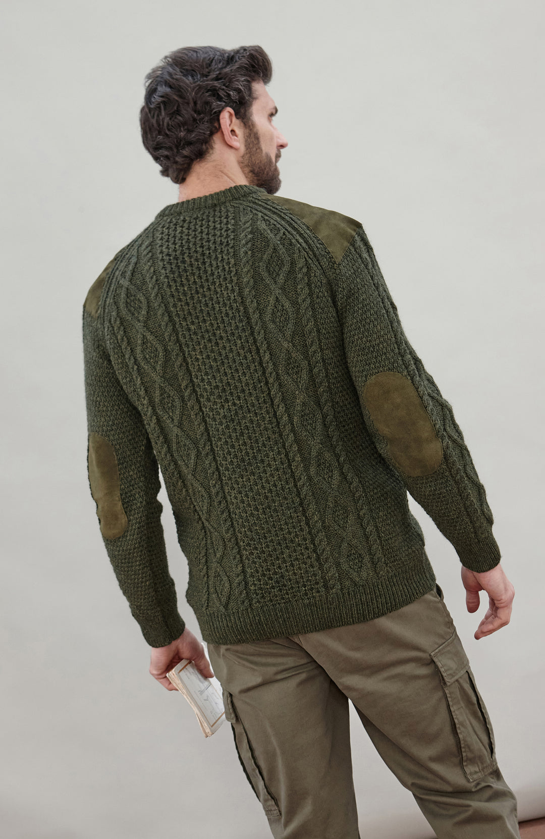 A man shown from the back wearing an olive green cable-knit sweater with brown elbow patches, paired with khaki cargo pants, against a neutral light gray background.